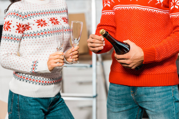 abgeschnittenes Bild eines Mannes, der eine Champagnerflasche öffnet, während seine Freundin mit Gläsern zur Feier des Umzugs im neuen Zuhause in der Nähe steht  - Foto, Bild