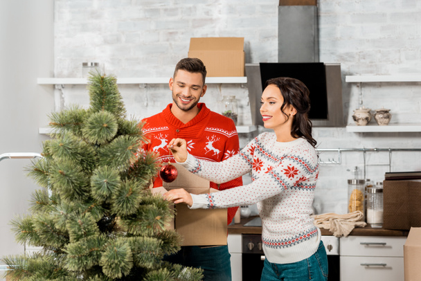 happy young couple decorating christmas tree by baubles in kitchen at home - Foto, Imagen