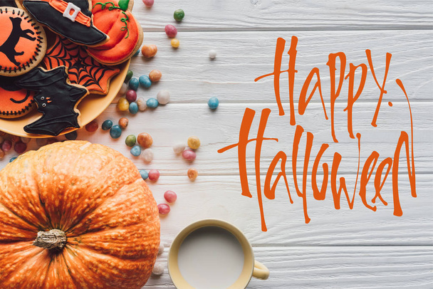 erhöhte Ansicht von Kürbis, Teller mit Halloween-Plätzchen, Bonbons und Tasse mit Milch auf Holzgrund mit "Happy Halloween" -Schriftzug - Foto, Bild