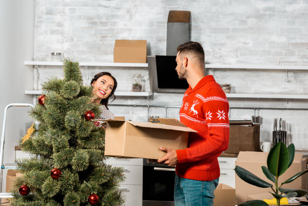 side view of man holding cardboard box while his girlfriend decorating christmas tree in kitchen at home  - Fotoğraf, Görsel