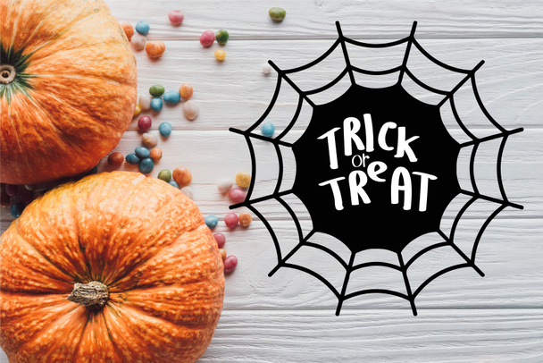 top view of pumpkins and colorful candies on wooden background with spider web and "trick or treat" lettering - Fotó, kép