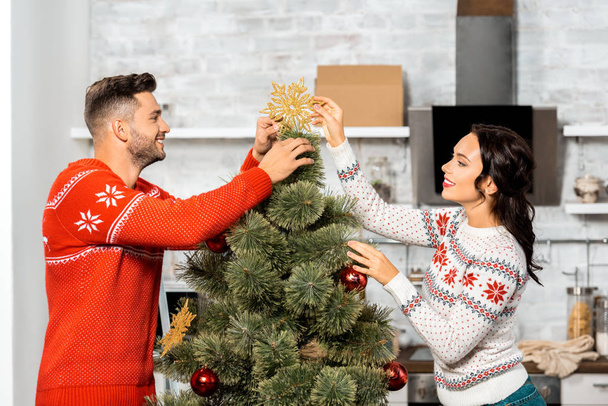 vista lateral de feliz pareja joven decorando el árbol de Navidad en casa
 - Foto, Imagen