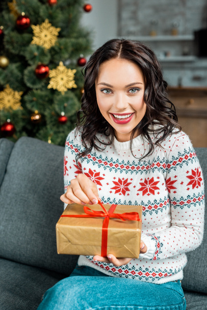 smiling young woman with gift box on sofa near christmas tree at home - Foto, afbeelding