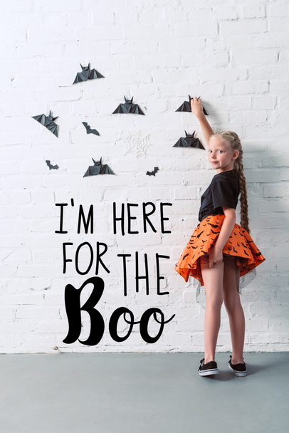 back view of child in skirt hanging black paper bats on white brick wall, halloween holiday concept with "Im here for the Boo" lettering - Photo, Image