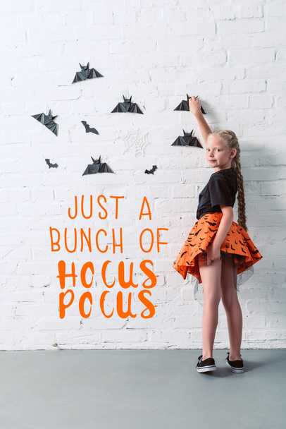 back view of child in skirt hanging black paper bats on white brick wall, halloween holiday concept with "just a bunch of hocus pocus" lettering - Photo, Image