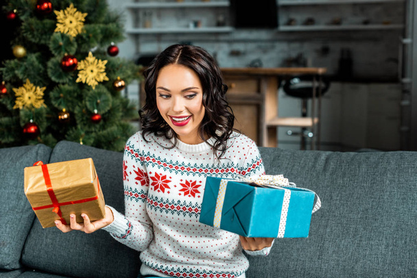 happy young woman holding gift boxes on sofa near christmas tree at home  - Фото, изображение