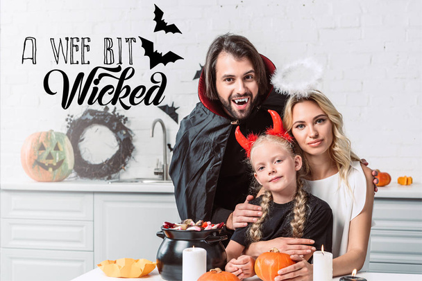 portrét rodičů a dcery v halloween kostýmy u stolu s dárky v černý hrnec v kuchyni doma s nápisem "trochu zlý" - Fotografie, Obrázek