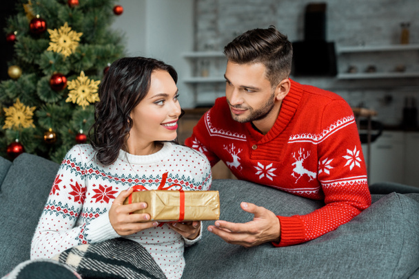 smiling young man gifting present box to attractive girlfriend on sofa near christmas tree at home - Foto, Imagen