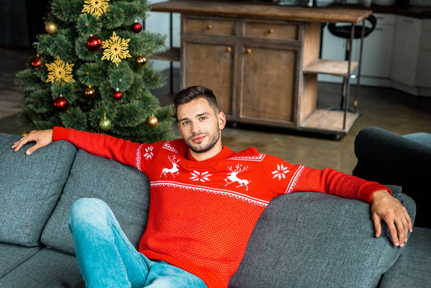fiatal jóképű férfi ül a kanapén, otthon a karácsonyfa közelében - Fotó, kép