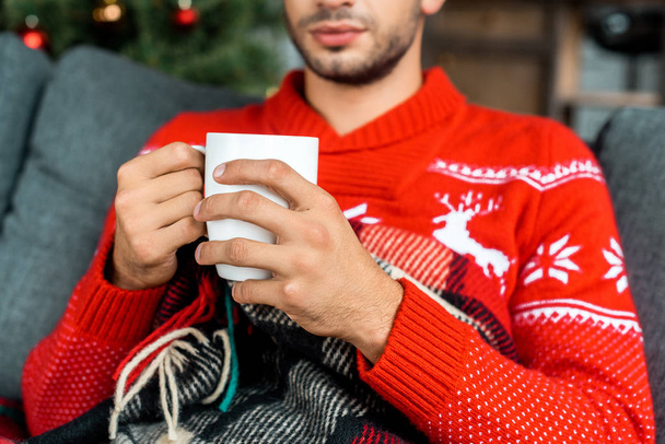 partial view of man sitting on sofa under blanket  holding cup of coffee near christmas tree at home - Foto, imagen