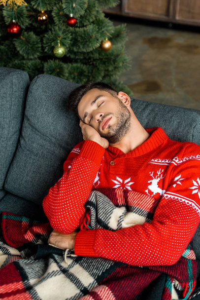 pohledný mladý muž spí na pohovce pod dekou u vánoční stromeček doma  - Fotografie, Obrázek