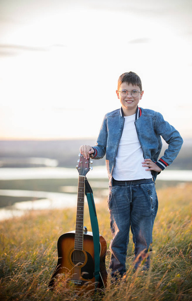Giovane ragazzo adolescente che tiene la chitarra acustica al campo estivo sul tramonto e sorridente
 - Foto, immagini