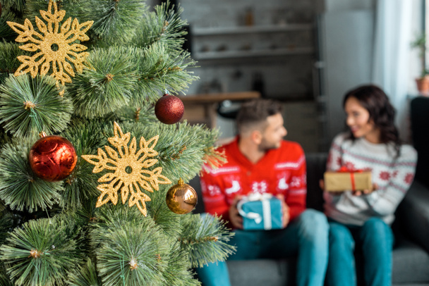 selective focus of decorated christmas tree and couple with gift boxes sitting on sofa behind at home  - Foto, afbeelding