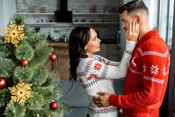 side view of couple embracing and looking at each other near christmas tree at home - Photo, Image