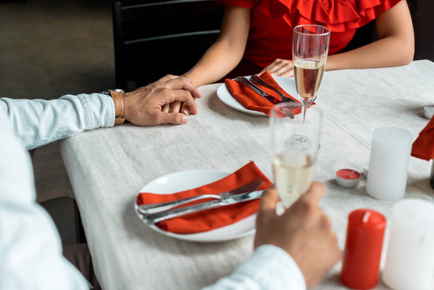 bijgesneden afbeelding van paar, houdend handen en het vieren van Kerstmis met champagneglazen aan geserveerd tafel met kaarsen thuis - Foto, afbeelding