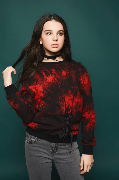 Young brunette teen model girl in trendy red-black sweatshirt, in grey jeans and in a black choker posing at dark-green background at studio - Photo, image