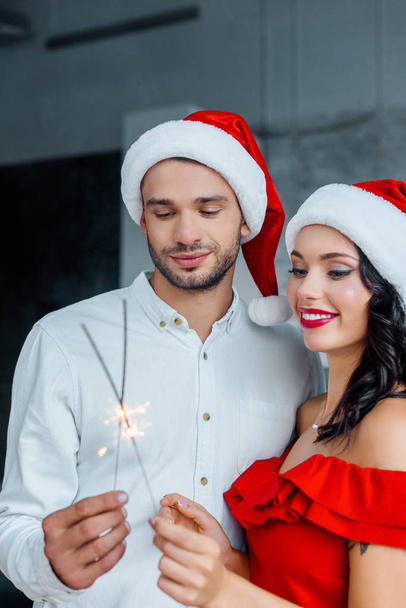 focus selettivo di giovani coppie in cappelli di Natale che celebrano con luci bengala a casa
 - Foto, immagini