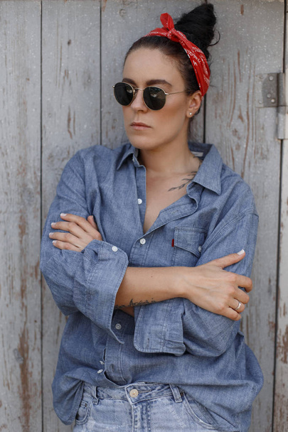Stylish beautiful woman with sunglasses in denim fashionable clothes and jeans with a bandana stands near a vintage wooden wall - Photo, Image