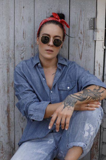 Belle femme hipster américaine élégante avec un tatouage sur les mains dans une chemise en denim de mode et un jean bleu vintage avec des lunettes de soleil se trouve près d'un mur en bois
 - Photo, image