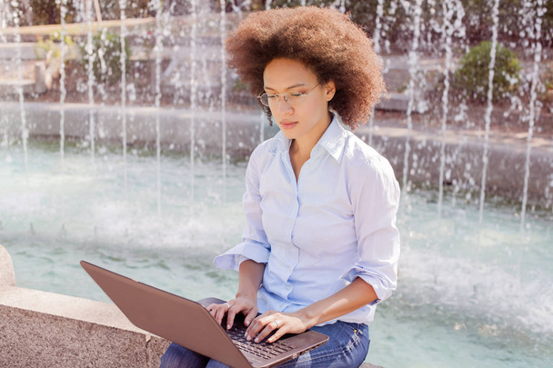 Kaunis nuori afroamerikkalainen opiskelija nainen, jolla on lasit ja joka työskentelee kannettavan tietokoneen kanssa. Ulkouima muotokuva, rento kuluminen, katsot tietokonetta
 - Valokuva, kuva