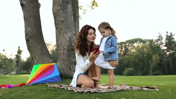 Mother and daughter outdoors. Little daughter holding smartphone. Weekend time in the park. - Video