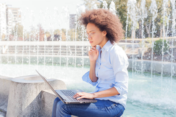 Mooie jonge Afro Amerikaanse Student vrouw met glazen werken met de Laptop. Outdoor Portret, vrijetijdskleding, kijken naar de Computer - Foto, afbeelding