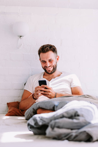 Молодой человек, использующий смартфон в постели утром
 - Фото, изображение