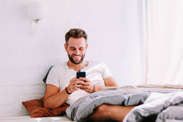 Νεαρός άνδρας χρησιμοποιώντας το smartphone στο κρεβάτι το πρωί - Φωτογραφία, εικόνα
