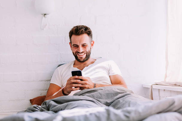 Νεαρός άνδρας χρησιμοποιώντας το smartphone στο κρεβάτι το πρωί - Φωτογραφία, εικόνα