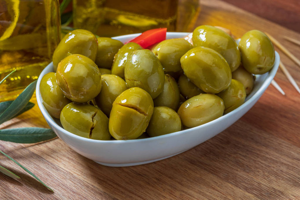 Artisan olives (canned in extra virgin olive oil, vinegar, spices) with red peppers and garlic. Includes olive tree leaves, slices of bread and bottle of extra virgin olive oil. Appetizer concept. On wooden board background. - Фото, изображение