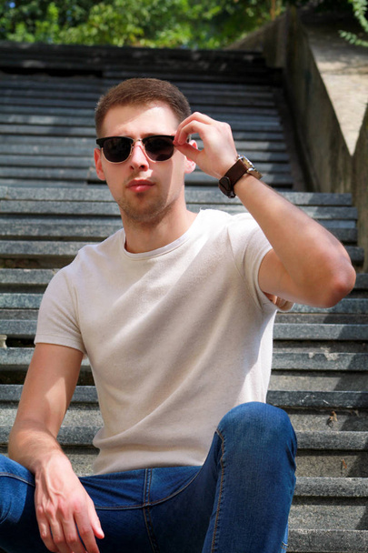 Man sitting alone on steps. Handsome boy with sunglasses. Male model posing for shooting, sitting on old stairs. Portrait of cool guy sitting on marble staircase waiting for someone or something. - Foto, Bild