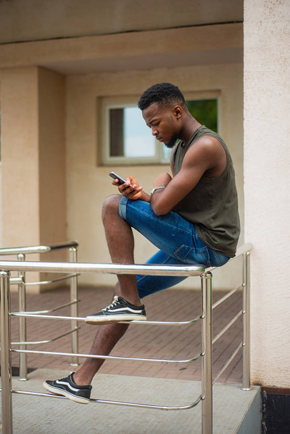 Hipster, a young man using a smartphone. African American teenager holding a mobile smartphone sitting on the railing - Foto, Imagen