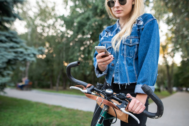 Stylish girl is parked with a bike and uses a smartphone. Sports and Technology. Woman with a bicycle is using the internet on a smartphone in the park. - Photo, Image