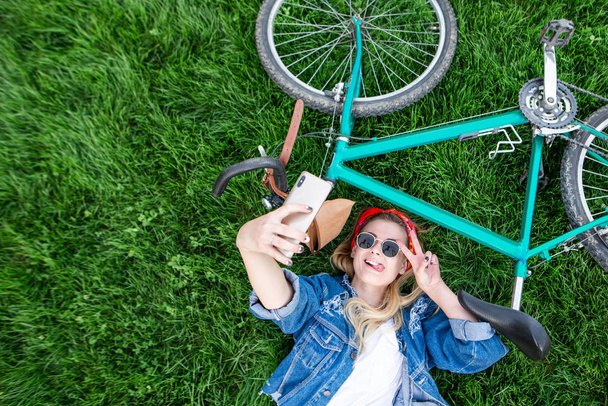 Módní ženu v džínové bundě a sluneční brýle si klade na trávě se na kole a bere selfie na smartphone. Stylová holka leží na trávníku s kol a dělá selfie. Copyspace - Fotografie, Obrázek