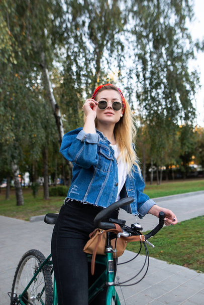 Portrait of a stylish young woman standing on a ride in a bike park and fixing glasses. Attractive girl with a bicycle walking around the park. Active leisure concept - Foto, Imagen