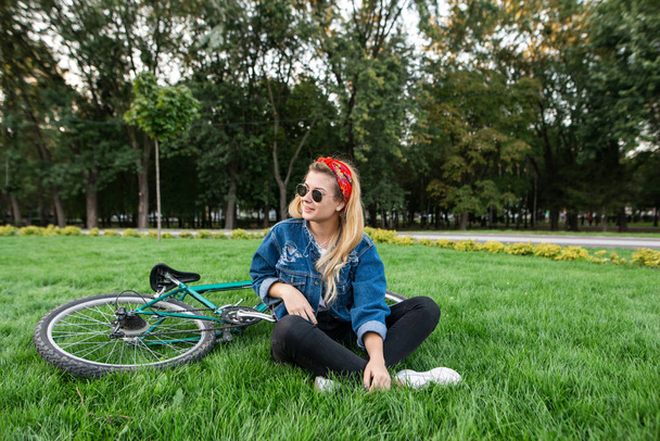 Stijlvolle aantrekkelijke jonge vrouw zit op een gazon in een park op een gazon en glimlacht. Mooi meisje in een stijlvolle outfit rusten op het gras tijdens het lopen op een fiets. Ontspannen buitenshuis - Foto, afbeelding