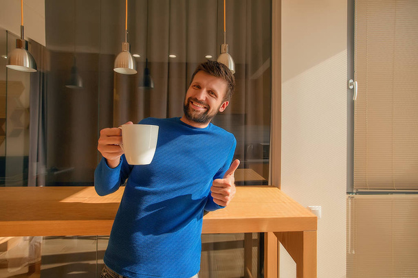 Lächelnder bärtiger Mann mit weißer Kaffeetasse und gestikulierendem Daumen im Innenraum - Foto, Bild