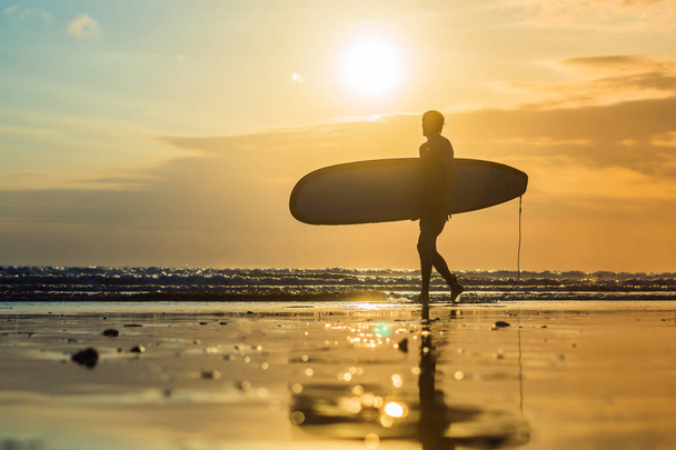 young man walking with surfboard along seashore at sunset - 写真・画像
