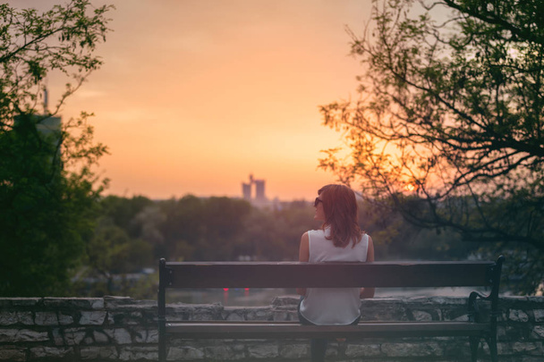 alleinstehendes Mädchen sitzt auf einer Bank und blickt in die ferne Stadtkulisse. - Foto, Bild