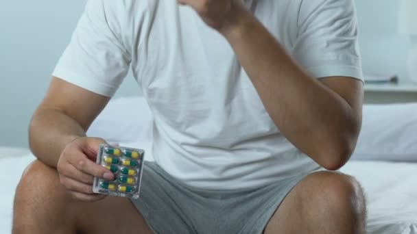 Sad handsome man sitting on bed holding pills blister, mens health problem - Felvétel, videó