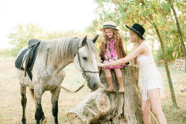 Beautiful young girl with blond hair in a suede jacket with fringe with little sister in a straw hat and checkered vintage dress with a horse in the countryside on a sunny autumn day - Photo, image