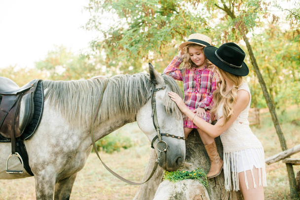 Beautiful young girl with blond hair in a suede jacket with fringe with little sister in a straw hat and checkered vintage dress with a horse in the countryside on a sunny autumn day - Фото, изображение
