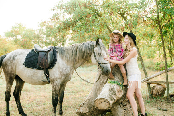 Beautiful young girl with blond hair in a suede jacket with fringe with little sister in a straw hat and checkered vintage dress with a horse in the countryside on a sunny autumn day - Photo, Image