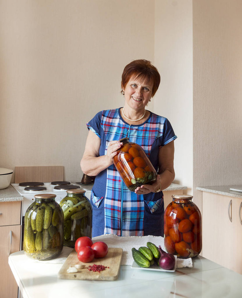 1 white adult woman with cans of pickled cucumbers and tomatoes in the kitchen - Foto, Imagen