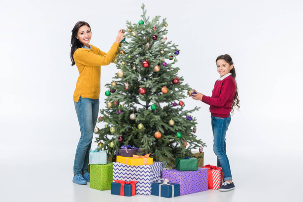 Madre feliz con hija decorando el árbol de Navidad con bolas de vidrio y mirando a la cámara aislada en blanco
 - Foto, Imagen