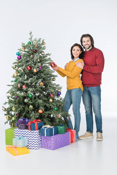 Feliz pareja decorando el árbol de Navidad con regalos y mirando a la cámara aislada en blanco
 - Foto, Imagen