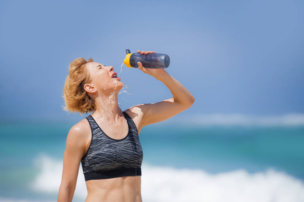 young happy and attractive sport runner woman drinking water bottle or isotonic drink after running workout at tropical paradise beach showing fit and athletic body in healthy lifestyle concept - Foto, imagen