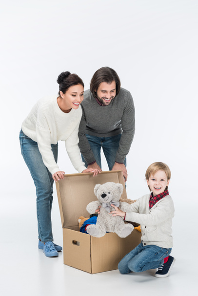 Padres felices con hijo sosteniendo oso de peluche y sentado cerca de la caja de cartón con juguetes aislados en blanco
  - Foto, Imagen