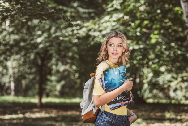 portrait of beautiful young woman with backpack and magazines in park - Fotó, kép