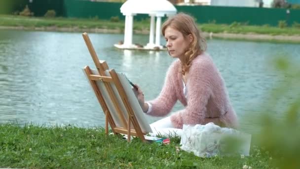 Μια όμορφη γυναίκα με τα κόκκινα μαλλιά, χρωματίζει μια εικόνα σε καμβά, που στέκεται στο καβαλέτο. Η κυρία είναι στο ύπαιθρο κοντά στην λίμνη του ποταμού, που αντλεί από τη ζωή - Πλάνα, βίντεο